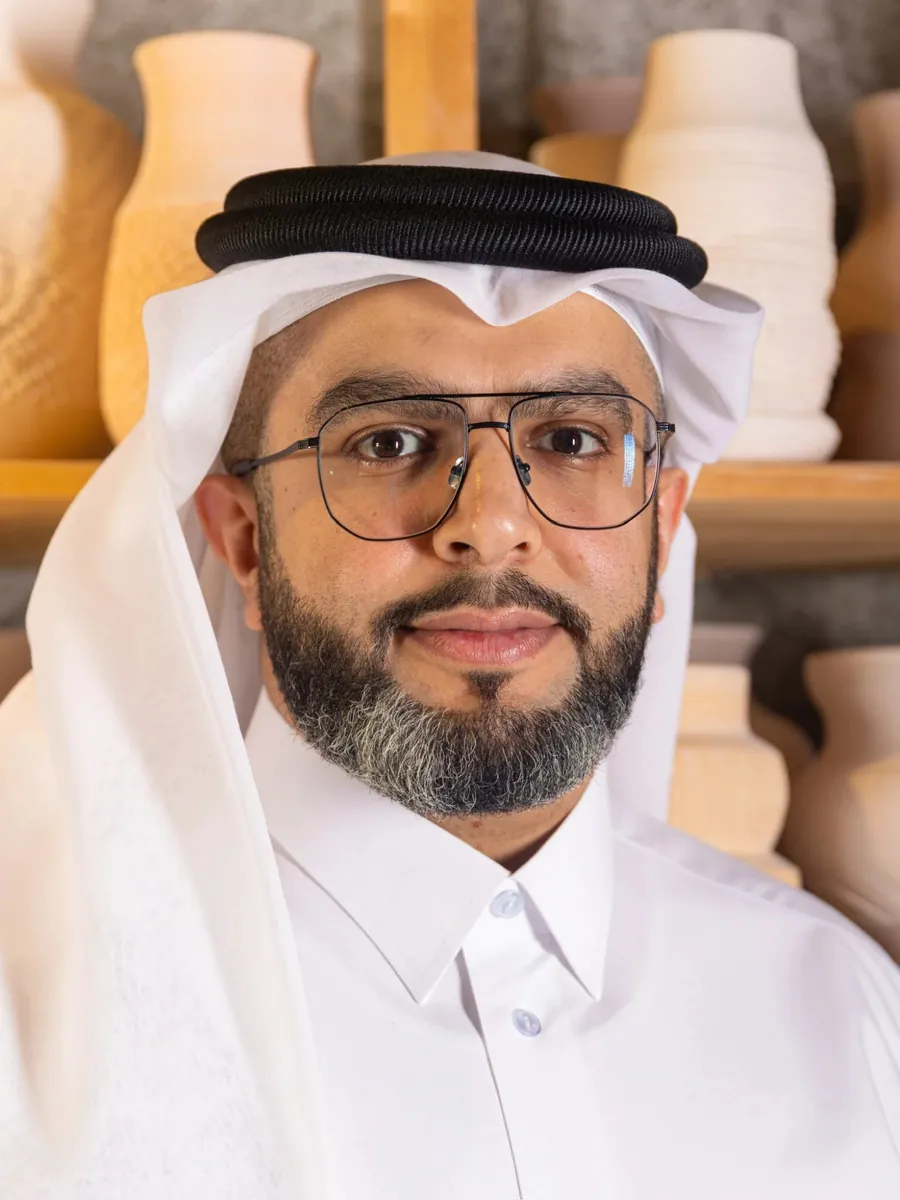Hameed Al-Qahtani in a white thobe and ghutra and dark-framed glasses, in front of shelves of unglazed clay vases.